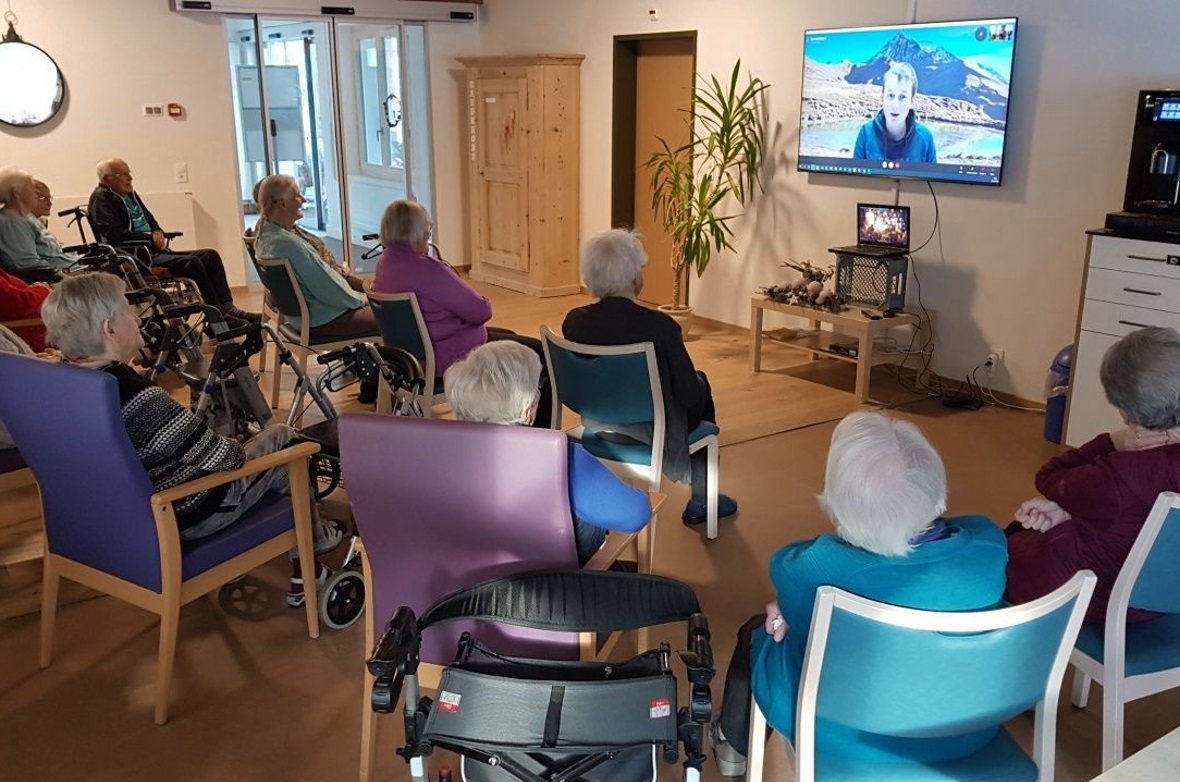 Manuel erscheint auf dem Bildschirm. Die Anwesenden hören aufmerksam zu.